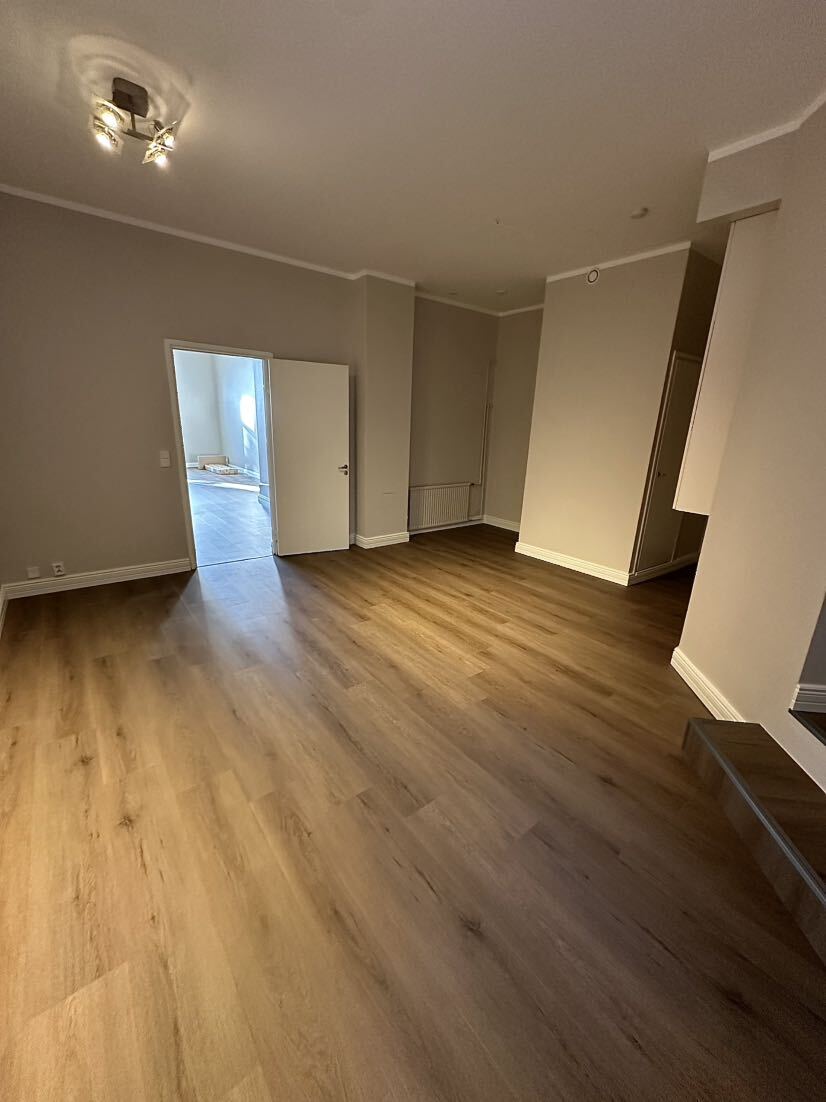 Tyhjä huone, jossa vaaleanruskea puulattia, vaaleat seinät ja avoin valkoinen ovi toiseen valoisaan huoneeseen.